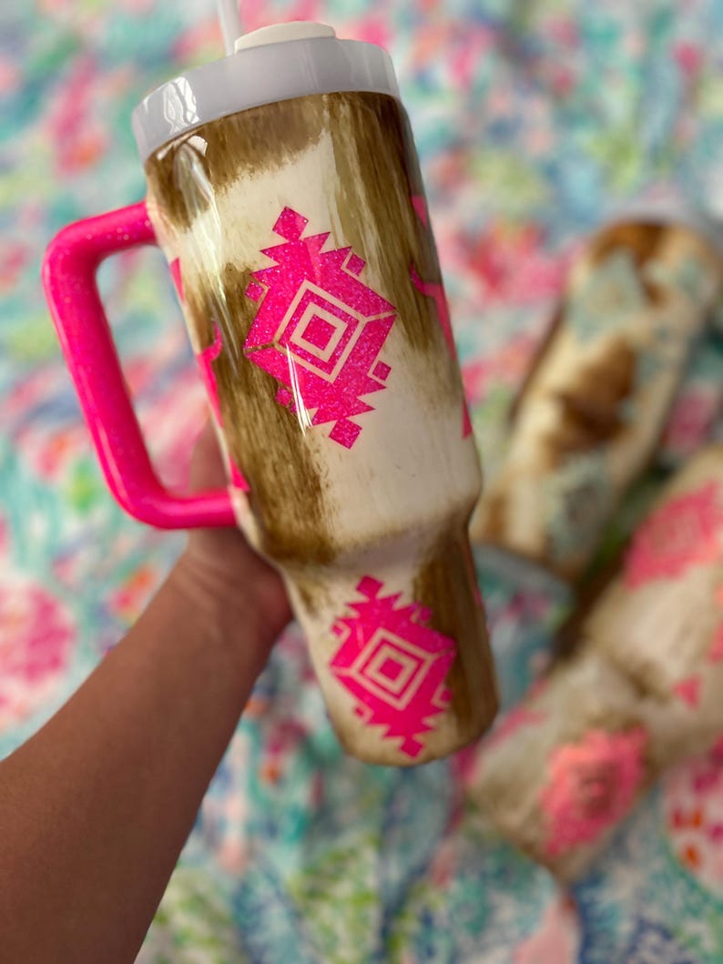 Puede incluir: Un vaso con un patr&oacute;n de piel de vaca marr&oacute;n y blanco y un asa rosa brillante. El vaso presenta un dise&ntilde;o geom&eacute;trico rosa. La tapa es blanca y se ve una pajita blanca. El vaso est&aacute; dise&ntilde;ado para contener una bebida.