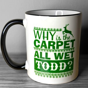 May include: White ceramic mug with a black handle. The mug has a green design with the text "WHY is the CARPET ALL WET TODD?" and a green reindeer graphic.