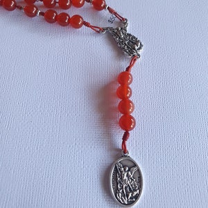 Puede incluir: Un rosario con cuentas de color rojo anaranjado y una medalla de San Miguel plateada. Las cuentas están ensartadas en un cordón rojo, con una pieza central decorativa plateada. El rosario se muestra sobre un fondo blanco.