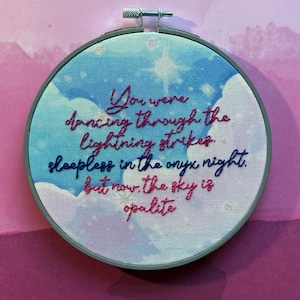 May include: Embroidered hoop art with a celestial theme. The design features a blue and white sky with clouds and a star. Text in pink and blue reads "You were dancing through the lightning strikes, sleepless in the onyx night, but now the sky is opalite."