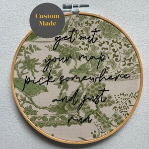 Pode incluir: Um aro de bordado redondo de cor creme com um padrão floral verde. O aro tem uma citação bordada em preto que diz: "get out your map, pick somewhere and just run."
