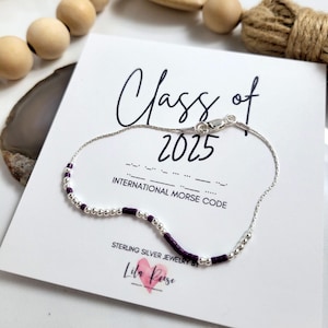 May include: A silver bracelet with purple beads and silver detailing. The bracelet is presented on a white card with the words "Class of 2025" and "International Morse Code". The card also has the text "Sterling Silver Jewelry By Lila Peise".
