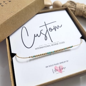 Peut inclure: Un bracelet en code Morse rempli d'or avec des perles dorées et turquoise, présenté dans une boîte en papier kraft. La carte à l'intérieur indique "Custom" et "International Morse Code". Les bijoux sont de Lila Reese.