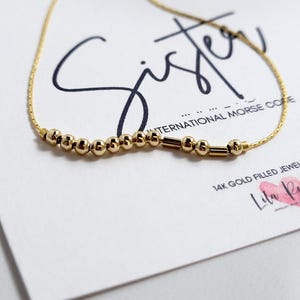 May include: A fine gold necklace featuring small, spherical beads and cylindrical bars, possibly spelling out a message in Morse code. The necklace is presented on a white card with the word "Sister" written in stylish script.