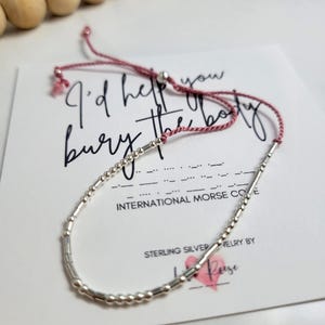 May include: A silver Morse code bracelet with a pink cord, displayed on a white card. The card features the text "I'd help you bury the body", alongside "International Morse Code" and "Sterling Silver Jewelry by La Rose".