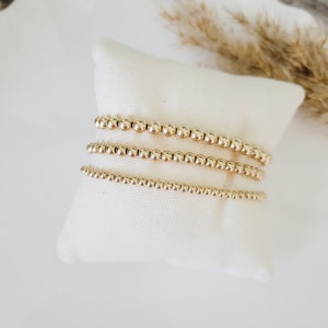 May include: Four gold beaded bracelets on a white cushion. The bracelets are made of small, round, gold beads. The cushion is on a white surface, with dried plants in the background.