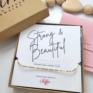May include: A gold-filled beaded necklace with the words "Strong & Beautiful" in Morse code, presented in a kraft paper box. The box also contains a pink envelope and a wooden heart-shaped bead. The jewelry is from Lila Reese.
