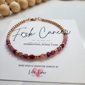 May include: Rose gold bracelet featuring pink gemstone beads and gold accents. The bracelet rests on a white card with the text "F*ck Cancer" and "International Morse Code". The card also has the text "Rose Gold & Gemstone Jewelry by Lila Reese".