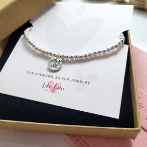 May include: A silver beaded bracelet with a round charm featuring the letter "B" inside a gift box. The bracelet is displayed on a white card with the text "925 STERLING SILVER JEWELRY" and the brand name "Lila Rose". The box is brown.