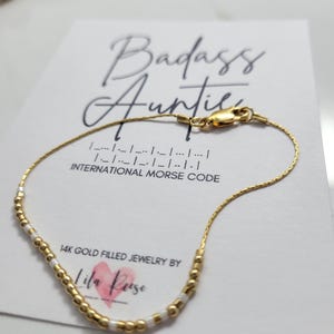 Peut inclure: Un bracelet en or rempli de perles blanches et dorées, présenté sur une carte avec les mots "Badass Auntie" et "Code Morse International". La carte comporte également le texte "14K Gold Filled Jewelry by Lila Rose."