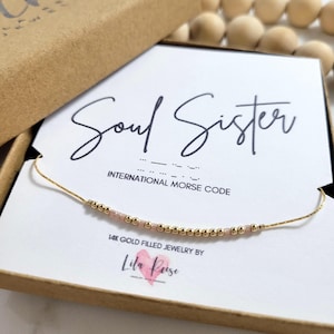 May include: A gold-filled necklace with pink and gold beads, presented in a brown gift box. The card reads "Soul Sister" in script, with Morse code symbols below. The necklace is from Lila Rose.