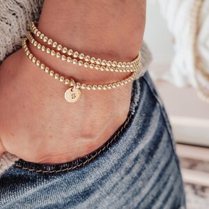 Peut inclure: Trois bracelets dorés à perles sont empilés sur un poignet. Les bracelets sont faits de petites perles rondes. Un bracelet a un petit charme rond avec la lettre "S". La personne porte un jean bleu.