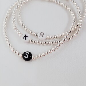 May include: Three silver beaded bracelets with lettered beads. One bracelet has a black bead with the letter "S", another has a white bead with the letter "R", and the third has a white bead with the letter "K".