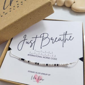 May include: A silver necklace with black and silver beads spelling out "Just Breathe" in Morse code. The necklace is presented on a white card inside a brown jewelry box. The box is branded with "Lr Reese Jewelry".