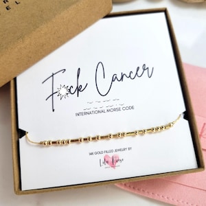May include: A gold-filled necklace with a Morse code design, presented in a gift box. The card inside reads "F*ck Cancer" with the words "International Morse Code" below. The box is from "Reese Jewelry".