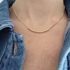 May include: A delicate gold necklace featuring small beads and a fine chain. The necklace is worn against a denim jacket, creating a contrast between the jewellery and the casual clothing.