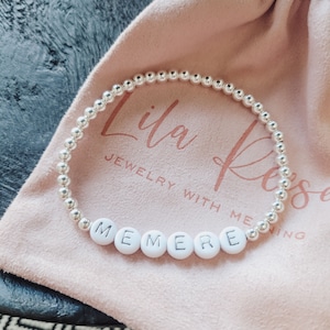 May include: A silver beaded bracelet with white letter beads spelling "MEMERE" rests on a pink cloth bag. The bag features the text "Lila Rose JEWELRY WITH MEANING" in a cursive font. The bracelet is the focal point of the image.