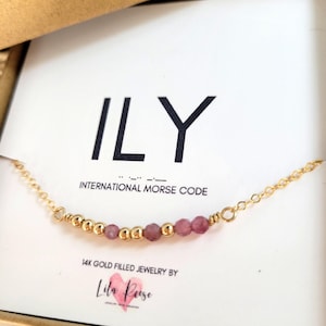 May include: A gold-filled necklace featuring pink beads and gold detailing. The necklace is displayed in a gift box with the text "ILY" and "International Morse Code". The brand name "Lila Peise" is also visible.