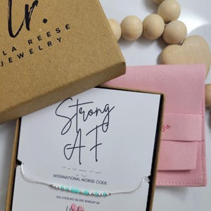 Peut inclure: Un collier en argent avec des perles turquoise dans une boîte cadeau. La boîte contient une carte avec les mots "Strong AF" et "International Morse Code". La boîte est marron et porte l'inscription "Lila Reese Jewelry".