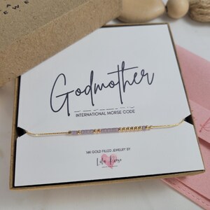 Peut inclure: Un collier en or rempli avec une chaîne délicate et de petites perles épelant "Godmother" en code Morse. Le collier est présenté sur une carte blanche à l'intérieur d'une boîte cadeau marron. La carte comprend également le texte "14K Gold Filled Jewelry by Lila Reese".