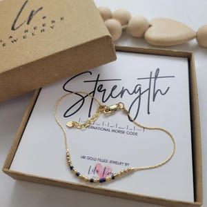 May include: A gold bracelet with navy blue beads, displayed in a brown box. The box contains a card with the word "Strength" and the text "14K Gold Filled Jewelry by Lila Reese". The bracelet is designed with the International Morse Code.