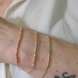 May include: Three delicate gold bracelets, each with small, colourful beads. One bracelet has coral beads, another has white and pink beads, and the third has turquoise beads. The bracelets are displayed on a person's arm.