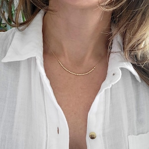 May include: A delicate gold necklace featuring small beads and bars arranged in a curved pattern. The necklace is worn against a white shirt, offering a minimalist and elegant aesthetic. The necklace is a subtle, yet stylish accessory, perfect for everyday wear.