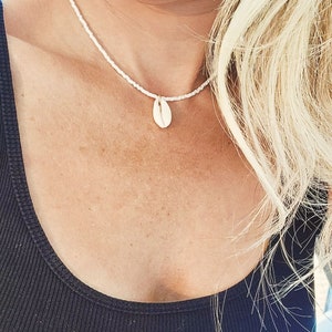 May include: A white beaded necklace featuring a cowrie shell pendant. The necklace is set against a black top, with blonde hair visible. The shell pendant is a natural, off-white colour.