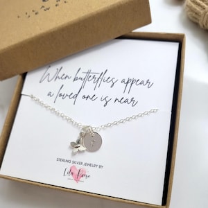 May include: A silver necklace with a small butterfly charm and a circular disc pendant engraved with the letter F. The necklace is displayed on a white card featuring the text When butterflies appear a loved one is near inside a brown box.