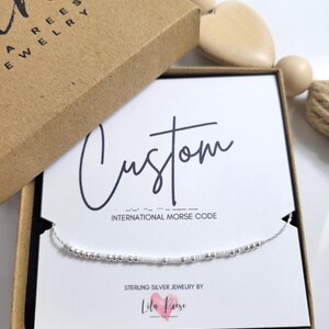Peut inclure: Un bracelet en argent avec un motif en code Morse est présenté sur une carte blanche à l'intérieur d'une boîte marron. La carte indique "Custom" en cursive et "International Morse Code". La boîte est étiquetée "Lila Reese Jewelry".