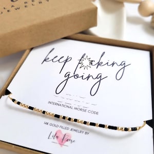 May include: A gold-filled necklace with black beads spelling out "keep going" in Morse code. The necklace is displayed on a white card inside a brown box. The box has the text "Lr Jewelry" on it.