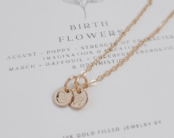 May include: A gold-filled necklace with two small, round pendants, one with a floral design and the other with a letter. The necklace is displayed on a card with the text "BIRTH FLOWERS" and the brand name "Lila Reize".