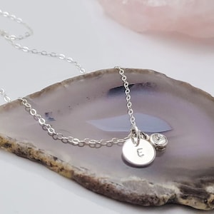 May include: A delicate silver necklace with a small round pendant engraved with the letter "E", alongside a clear gemstone. The necklace is displayed on a grey and white surface.