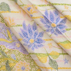 Könnte beinhalten: Eine Nahaufnahme eines weißen Stoffes mit einem Blumenmuster. Der Stoff zeigt ein sich wiederholendes Muster aus lila, gelben und grünen Blumen auf weißem Hintergrund. Der Stoff ist in der Mitte gefaltet, sodass das Muster auf beiden Seiten sichtbar ist.