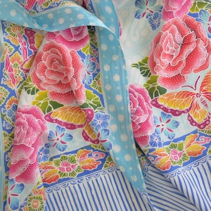 Può includere: Un foulard di seta blu e bianco con un motivo floreale. Il foulard presenta rose rosa, farfalle e altri fiori. I bordi del foulard sono rifiniti con un bordo a righe blu e bianco.