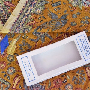 Könnte beinhalten: Eine weiße Box mit einem durchsichtigen Fenster, beschriftet mit "TJOA GIOK TJIAM" in blauer Schrift. Die Box liegt auf einem braunen Stoff mit einem blauen und weißen Blumenmuster.