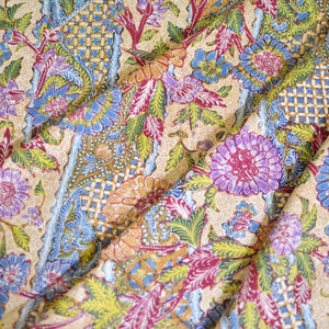 Può includere: Un tessuto beige con un motivo floreale colorato. Il tessuto presenta un motivo ripetitivo di fiori e foglie blu, rossi, gialli e verdi. Il tessuto è piegato in modo da mostrare il motivo.