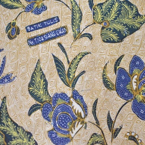 May include: A beige fabric with a blue and yellow floral pattern. The fabric has a repeating pattern of flowers and leaves. The fabric has the text "Batik Tulis" and "Nw. Tioa Siang Gwan" printed on it.