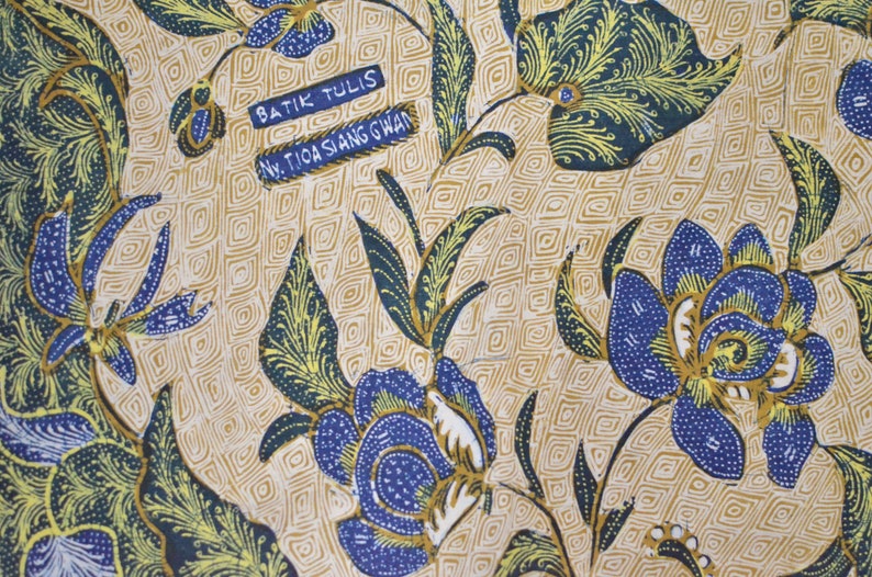 May include: A beige fabric with a blue and yellow floral pattern. The fabric has a repeating pattern of flowers and leaves. The fabric has the text "Batik Tulis" and "Nw. Tioa Siang Gwan" printed on it.