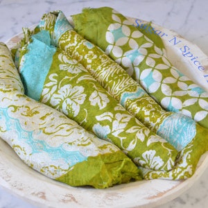 May include: Four green and white patterned fabric napkins folded and stacked in a white wooden bowl. The napkins have a floral design and are made of a soft, lightweight fabric. The bowl is round and has a rustic, distressed finish.