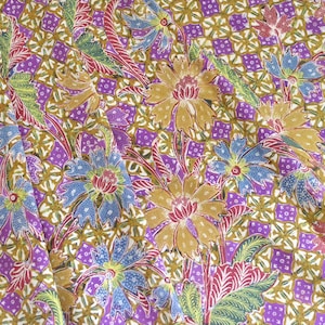 Könnte beinhalten: Nahaufnahme eines Stoffes mit floralem Muster. Das Design zeigt große Blüten in Gold-, Blau- und Rottönen vor einem violetten Hintergrund. Der Hintergrund hat ein geometrisches Muster. Der Stoff scheint ein Batik oder ein ähnliches Textil zu sein.