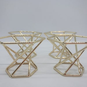 May include: Four gold-colored metal makeup sponge holders. Each holder is shaped like a hexagon with a geometric design.
