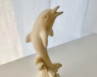 May include: A white porcelain dolphin figurine leaping out of the water. The dolphin is painted with gold accents and is set on a base with waves.
