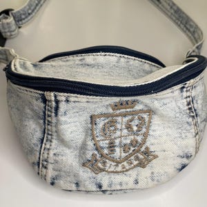 May include: A light blue denim fanny pack with a black zipper and a brown embroidered crest. The crest features a crown and the words "New York City".