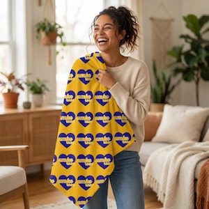 May include: A yellow blanket featuring a pattern of blue hearts and the text "umke too". The blanket is held by a person wearing a cream sweater and blue jeans. The background shows a living room with plants and furniture.
