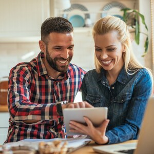 May include: A smiling couple looking at a tablet. The man wears a red and black plaid shirt, and the woman wears a denim jacket. They are indoors, possibly in a kitchen, with a laptop and papers visible on the table.