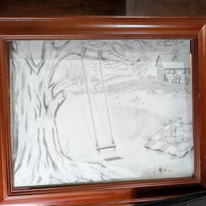 May include: A framed pencil drawing of a rural scene. The artwork depicts a large tree with a swing, a stone wall, and a house in the background. The frame is made of dark brown wood.