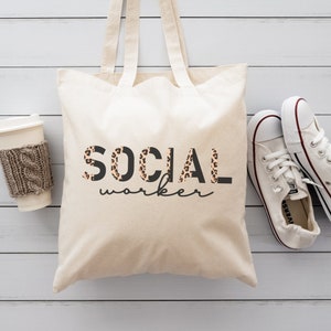 Puede incluir: Bolso tote color crema con las palabras "SOCIAL worker" impresas en negro y estampado de leopardo. El bolso tiene dos asas y está junto a un par de zapatillas blancas y una taza de café con una funda de punto.
