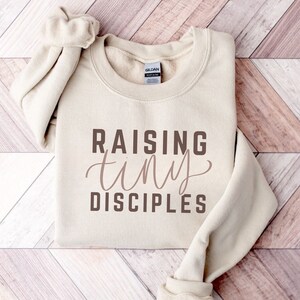 May include: A beige sweatshirt with the text "Raising tiny disciples" printed in brown on the front.