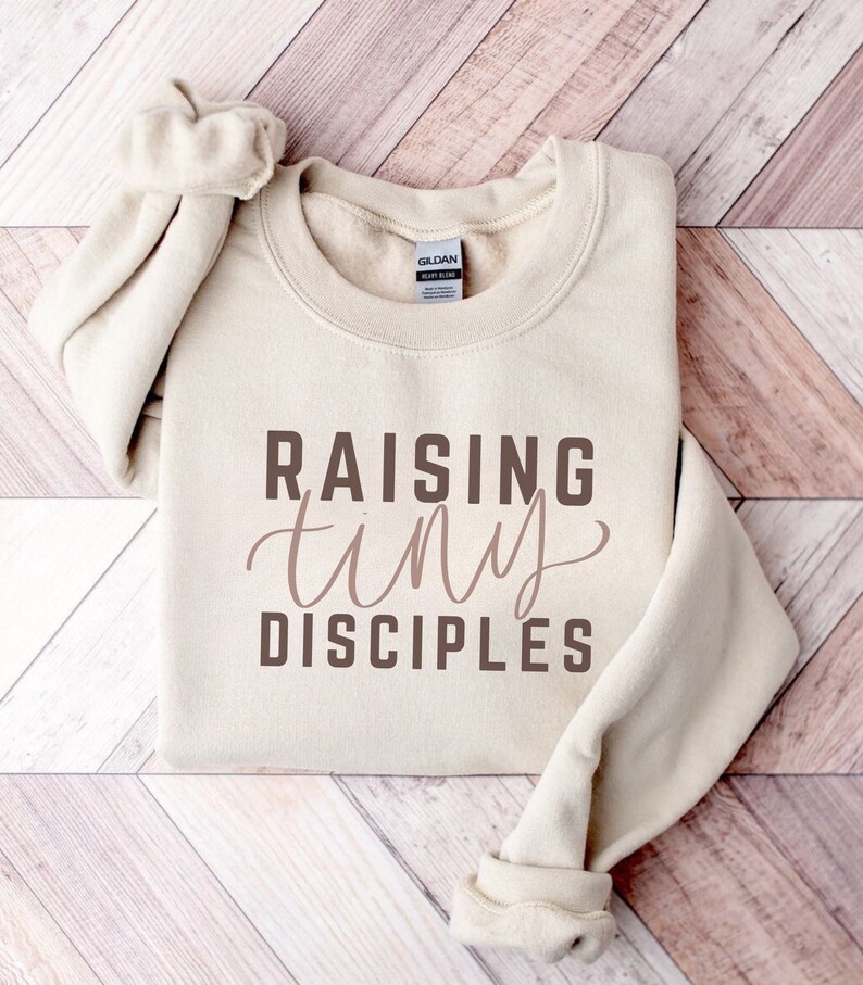 May include: A beige sweatshirt with the text "Raising tiny disciples" printed in brown on the front.